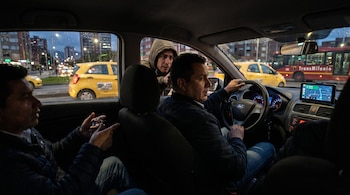 Vista interior de un coche en la ciudad al anochecer, con un hombre encapuchado asomándose por la ventanilla del conductor y un pasajero en primer plano.