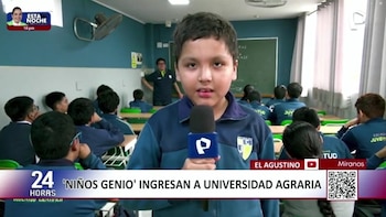 Primer plano de un niño hablando a un micrófono en un aula escolar. Detrás de él, varios estudiantes y un maestro. Pizarrón, pupitres y ventilador son visibles