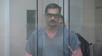 Primer plano de Jesse Lee Calhoun, un hombre con gafas, bigote y camiseta azul sobre una rosa, detrás de un cristal protector en una sala