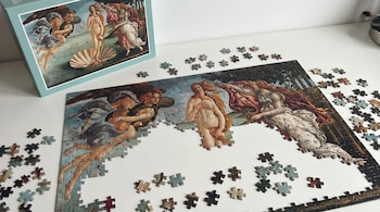Rompecabezas a medio armar de la pintura "El Nacimiento de Venus" de Botticelli, con piezas sueltas y la caja original sobre una mesa blanca.