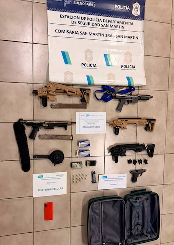 Seis armas de fuego, municiones, un teléfono celular rojo y una maleta abierta de color verde sobre baldosas, con un banner policial al fondo