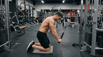 Hombre musculoso sin camisa arrodillado en una colchoneta, agarrando una cuerda de una máquina de polea alta en un gimnasio.