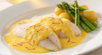 Dos filetes de merluza blanca con salsa amarilla de azafrán, hebras rojas, patatas pequeñas y espárragos verdes en un plato blanco.