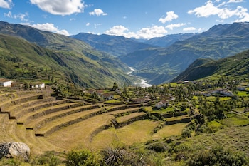 Terrazas agrícolas en semicírculo, casas blancas con tejados rojos, montañas verdes, un río en el fondo y cielo azul con nubes.