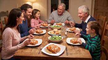 Seis personas, adultos y niños, comen en una mesa de madera con platos de pastel de zanahoria y carne, ensalada, pan y agua.