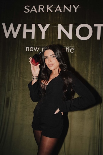 Mujer de cabello oscuro y traje corto negro sostiene una botella de perfume rojo y negro. Al fondo, un banner verde con texto blanco 'SARKANY WHY NOT'
