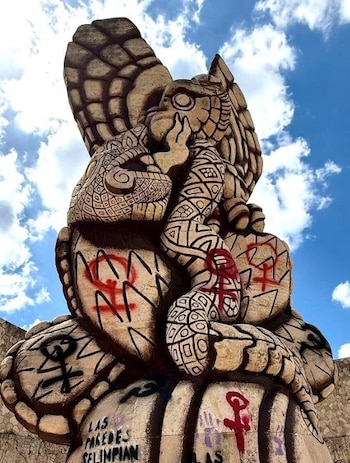 Escultura de águila del Monumento a la Patria en Mérida, con grafitis feministas rojos y negros. Se observa parte de la estructura base y cielo azul nublado de fondo