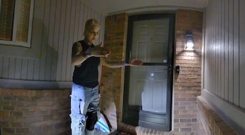 Una mujer rubia con tatuajes en los brazos, usando top oscuro y jeans rotos, gesticula con las manos abiertas frente a una puerta en un porche iluminado