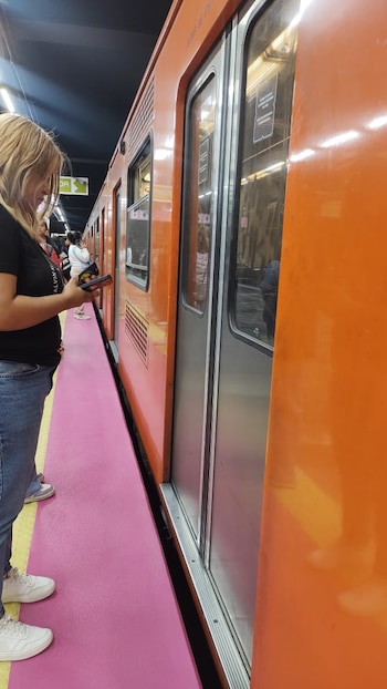 Una mujer de cabello rubio, con jeans y camiseta negra, de pie en una plataforma rosa de una estación de metro, junto a un tren naranja