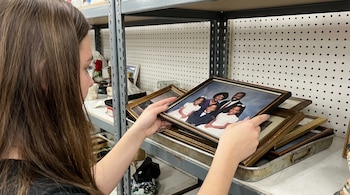 Una joven de perfil con cabello castaño oscuro sostiene un retrato enmarcado de una familia afroamericana elegantemente vestida en una tienda de segunda mano.