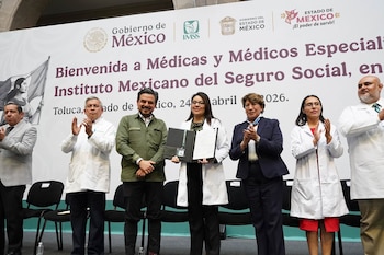 Un grupo de seis personas, incluidos cuatro en batas blancas, aplauden en un escenario frente a un gran banner del Gobierno de México y el IMSS