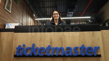 Una mujer con gafas sonríe detrás de un mostrador de madera con el logo azul de Ticketmaster. Al fondo se ve una pared de listones y barras metálicas
