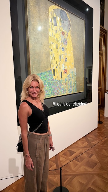 Yanina Latorre sonríe de pie frente a la gran pintura "El Beso" de Gustav Klimt en un museo. Lleva un top negro y pantalones anchos