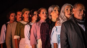 Ocho actores de diversas edades y géneros posan alineados en primer plano, mirando al frente, con un fondo oscuro y una iluminación frontal