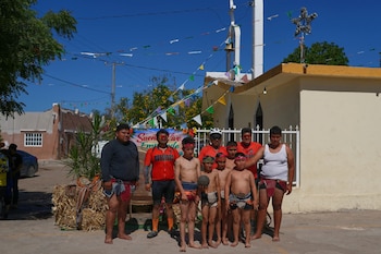 Un grupo de hombres y niños, algunos en atuendos tradicionales y un niño con un balón, posa al aire libre en México frente a un edificio y decoraciones festivas