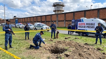 Agentes del INPEC y policía, perro K9, vehículos policiales, agujero en el suelo, tierra, pasto, muro de prisión, torre de vigilancia, cinta amarilla.