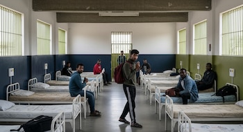 Gran sala de centro de migrantes con literas metálicas. Personas sentadas y caminando entre camas, paredes blancas, rejas y ventanas enrejadas visibles.