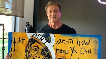 Un hombre sostiene una pintura rectangular que muestra el rostro estilizado de un hombre con sombrero y texto manuscrito sobre un fondo amarillo