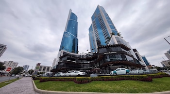 Dos rascacielos de cristal azul, un edificio comercial curvo oscuro, una avenida, varios vehículos, arbustos y césped.