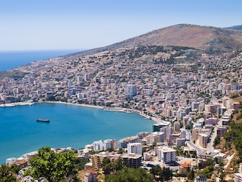 Vista aérea de una ciudad costera con múltiples edificios residenciales, una bahía de aguas azules, una playa, un barco, montañas y cielo