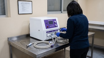 Una persona de espaldas con guantes azules examina los cables de un equipo blanco y violeta sobre una mesa metálica.