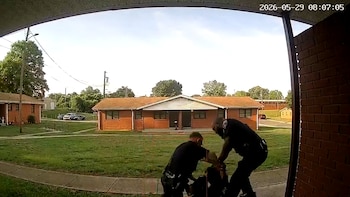 Dos policías uniformados sujetan a una persona en el suelo sobre una acera. Al fondo, se observan casas de ladrillo rojo y áreas verdes bajo un cielo claro