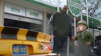 Shaquille O'Neal junto a un personaje militar animado frente a un taller de reparación y la parte trasera de un automóvil amarillo