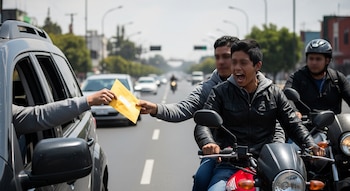 Brazo extendido desde ventana de coche sujeta sobre amarillo. Joven en motocicleta se estira para tomarlo, con la boca abierta, otro hombre lo acompaña.