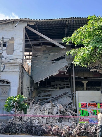 Edificio de dos plantas con pared parcialmente derrumbada, escombros en el suelo y cinta de seguridad. Se ven cables eléctricos, un cartel publicitario y vegetación