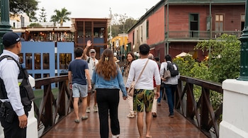 Varias personas caminan sobre un puente de madera con barandillas oscuras entre edificios de colores y un agente de seguridad en el extremo