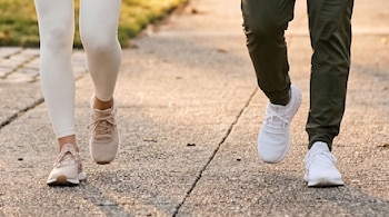 Piernas de dos personas, una con mallas blancas y zapatillas beige, la otra con pantalones verdes y zapatillas blancas, caminando en pavimento.