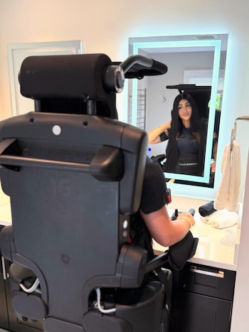 Una mujer de cabello largo vista desde atrás utiliza un dispositivo robótico negro frente a un espejo iluminado