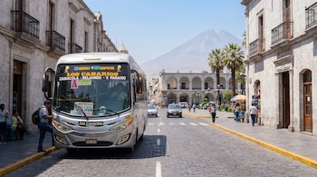 Bus plateado con pasajero abordando en calle adoquinada de Arequipa, edificios coloniales de sillar a los lados y volcán Misti en el horizonte.