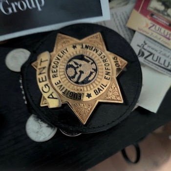 Un distintivo de metal dorado con forma de estrella, con las inscripciones 'AGENT', 'FUGITIVE RECOVERY' y 'BAIL ENFORCEMENT', sobre cuero negro con monedas