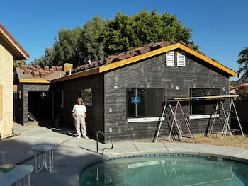 Casa en construcción con fachada cubierta de material negro, ventanas, tejas apiladas en el tejado. Andamios, una persona, piscina y patio con muebles