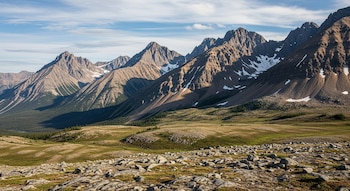 Paisaje montañoso de las Montañas Mackenzie con picos rocosos, laderas nevadas, valles verdes y un primer plano de rocas, bajo un cielo azul con nubes.