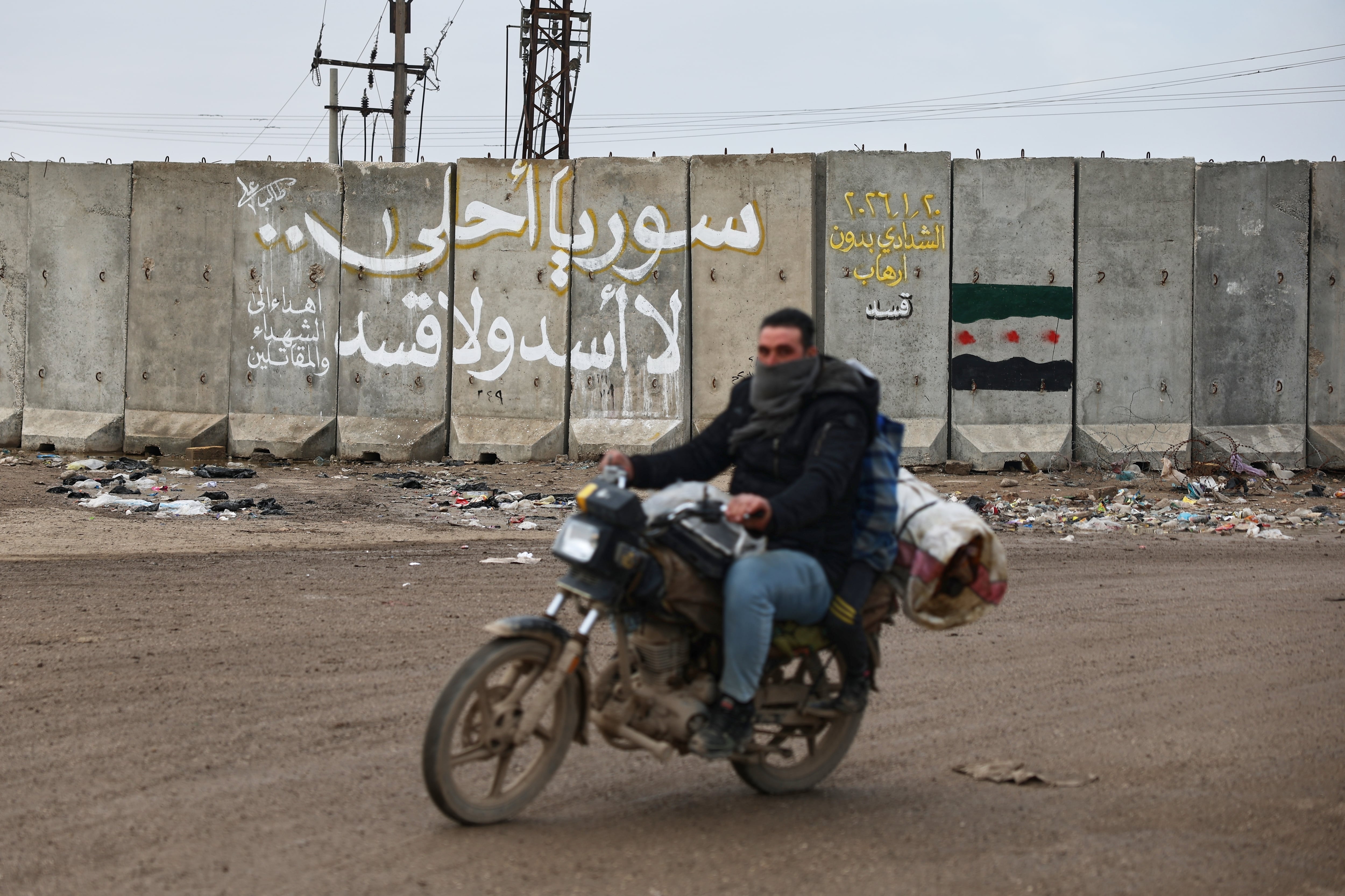 Un hombre en motocicleta pasa junto al muro de una base militar estadounidense con un grafiti en árabe que dice: