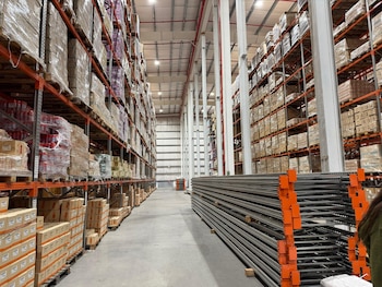 Interior de un almacén industrial con estanterías altas repletas de cajas en palés. En el suelo, a la derecha, hay vigas metálicas apiladas para construcción