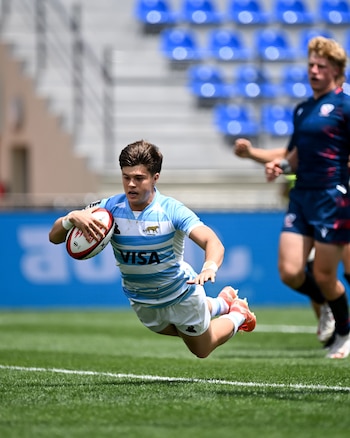 Jugador de rugby con camiseta celeste y blanca en plena zambullida sobre el césped con un balón ovalado. Otro jugador y asientos de estadio detrás
