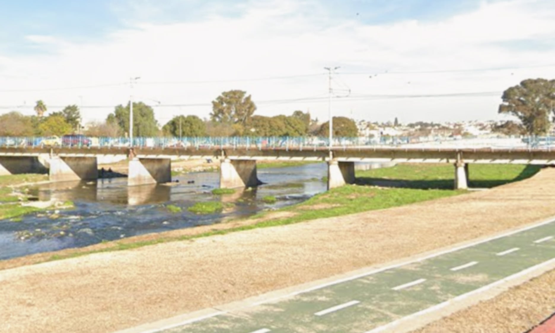 Un hombre se encuentra junto a un pilar del puente que cruza el río Suquía en la ciudad de Córdoba, con una ciclovía a la derecha del cauce (Google Maps)