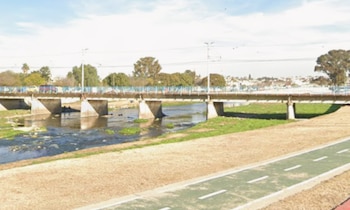 Puente sobre río con cauce bajo, orillas con vegetación seca y una ciclovía verde. Una figura humana borrosa se ubica junto a un pilar del puente