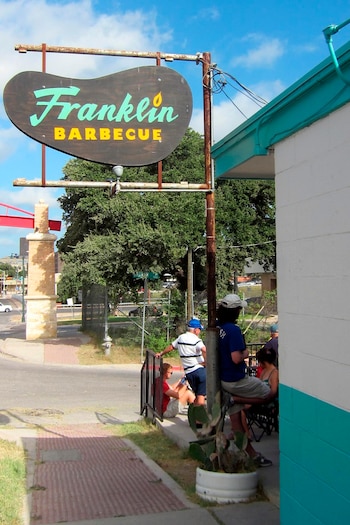 Vista exterior de Franklin Barbecue en Texas con su cartel marrón y turquesa, personas esperando afuera y parte de la fachada azul y blanca