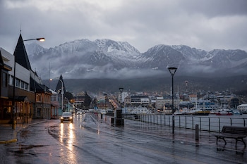 Calle mojada con reflejos de luces de autos y farolas, edificios costeros, un puerto con barcos y montañas con nieve y niebla al fondo.
