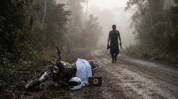 Motocicleta caída, casco médico blanco y botiquín rojo con cruz en una carretera de tierra, con densa vegetación y neblina. Una figura camina al fondo.