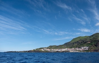 Mar azul frente a un poblado de casas blancas con tejados rojos en la falda de una colina verde, bajo un cielo azul con nubes tenues