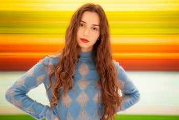 Una mujer con cabello largo ondulado y suéter azul posa con las manos en la cintura frente a un fondo con franjas de colores