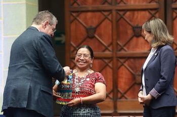 Un hombre de traje oscuro entrega un objeto colorido a una mujer con gafas y vestimenta tradicional, sonriente, mientras otra mujer observa