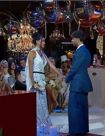 Una mujer con vestido blanco de novia y un hombre con traje azul se miran. Hay velas, lámparas de araña y esferas de espejo; varias personas observan