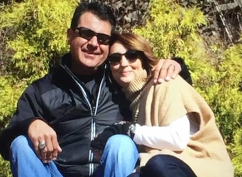 Un hombre y una mujer sonríen sentados al aire libre con gafas de sol y ropa de abrigo frente a vegetación
