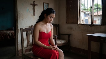 Una joven con cabello oscuro y vestido rojo se sienta en una silla de madera. Detrás, una pared texturizada con un crucifijo y una ventana enrejada.